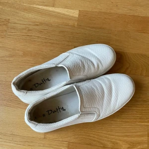 Vita slip-on sneakers från Duffy - Säljer ett par vita slip-on sneakers från Duffy med diskret ormmönstrad struktur. Skorna har elastiska paneler på sidorna för enkel på- och avtagning och en platt sula. Perfekta för en stilren och enkel look.