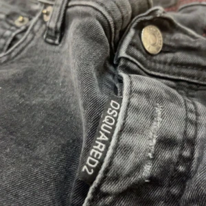 Svarta jeans från Dsquared2 - Säljer ett par svarta Dsquared2 jeans med diskreta slitningar och röda sömmar på insidan. Klassisk femficksmodell med knappgylf och Dsquared2-logga vid fickan. Snygg passform och coola detaljer. Size-s
