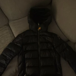 Svart dunjacka från Parajumpers - Säljer en svart dunjacka från Parajumpers, modell Sheen. Jackan har huva, dragkedja och klassisk pufferdesign med märkets logga på ärmen. Perfekt för kyliga dagar och riktigt snygg till vardags.