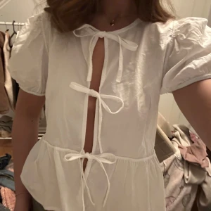 Vit blus med knytband - Säljer en söt vit blus med puffärmar och tre knytband framtill. Blusen har en luftig passform och volang nertill, perfekt för varmare dagar. Enkel och stilren design som passar till mycket.