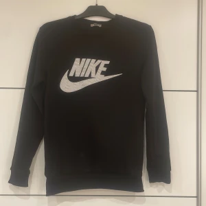 Svart sweatshirt från Nike med logga - Svart sweatshirt från Nike med stor vit logga och mönstrad text på bröstet. Tröjan har rund halsringning och långa ärmar. Perfekt för en sportig och avslappnad stil. Kan passa XS