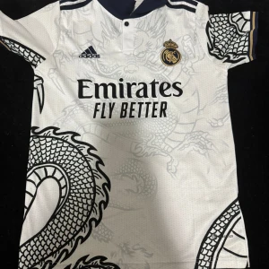 Real Madrid fotbollströja med drakmönster - Vit Real Madrid fotbollströja från Adidas med svart och guld detaljering. Stort drakmönster i grått över hela tröjan, klubbmärke och sponsortryck på bröstet. Kortärmad modell med sportig känsla och klassisk krage.