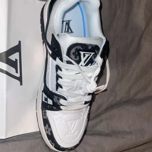Finns i alla storlekar  - Säljer ett par vita och svarta Louis Vuitton sneakers med klassiskt monogrammönster på detaljerna. Skorna har rund tå, platt sula och snörning. Märkeslogga syns tydligt på plösen och sidan. Perfekta för dig som vill sticka ut med lyxiga detaljer.