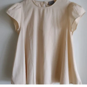 Beige a-linjeformad flowy blus med kort ärm - Säljer en beige blus med korta ärmar och rund halsringning. Blusen har en lös passform och en liten öppning med knapp i nacken. Perfekt för vår och sommar.