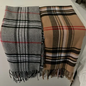 rutiga halsdukar - Säljer två snygga halsdukar med klassiskt rutigt mönster och fransar i kanten. En i grått med svarta, vita och röda ränder, och en i beige med svarta, vita och röda ränder. Perfekta accessoarer för kyliga dagar.