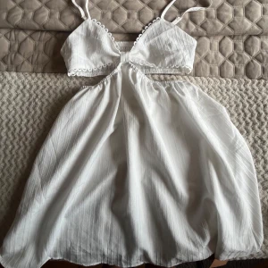Vit sommarklänning med cut-out och spetsdetaljer - Perfekt Söt vit studentklänning med smala axelband, cut-out under bysten och fina spetsdetaljer längs kanterna. Kjolen är lätt och luftig, perfekt för varma dagar. Klänningen har en feminin och ungdomlig stil.
