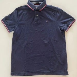 Marinblå piké från Tommy Hilfiger - Säljer en klassisk marinblå pikétröja från Tommy Hilfiger med korta ärmar. Kragen och ärmsluten har snygga ränder i vitt och rött. Liten broderad logga på bröstet och två knappar vid halsen. Perfekt för en stilren look!