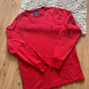 Röd long sleeve tröja från Polo Ralph Lauren - Säljer en röd långärmad tröja från Polo Ralph Lauren med klassisk blå logga broderad på bröstet. Tröjan har rund halsringning och är tillverkad i ett strukturerat bomullsmaterial. Kan nog både användas som sovplagg eller vardagligt. 
