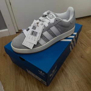 Grå Adidas Campus 00s sneakers (oanvända) - Säljer ett par nya gråa Adidas Campus sneakers med vita ränder och vit sula. Skorna har klassisk design med snörning och är tillverkade i mocka. Perfekta för dig som gillar stilrena och bekväma sneakers. Storlek 43 1/3. Köpta på Nelly Man.