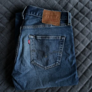 Blå jeans från Levi's - Säljer ett par klassiska blå jeans från Levi's med en snygg tvätt och femficksdesign. De har en normal passform och är perfekta för en avslappnad stil. Märkeslappen i brunt läder sitter på baksidan av midjan. Pris 199