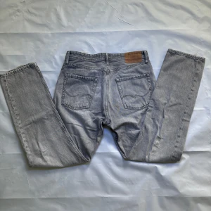Grå jeans från Jack & Jones - Säljer ett par grå jeans från Jack & Jones med klassisk femficksdesign och läderpatch bak i midjan. Jeansen har raka ben och normal passform, tillverkade i denim med lätt tvättad look. Har synliga hål, ett i fickan och två jättesmå på knäet.