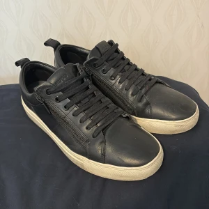 Hugo skor - Säljer ett par svarta sneakers från Hugo med ovandel i skinn och vita platta sulor. Skorna har snörning och diskret Hugo-logga på insidan och sulan. Perfekta för en stilren och clean look.