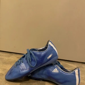 Blå Adidas fotbollsskor med snörning - Säljer ett par blå Adidas fotbollsskor med snörning och vit logga på sidan. Skorna har en låg profil och är tillverkade i syntetmaterial med mönstrad yta. Perfekta för dig som vill sticka ut på planen!