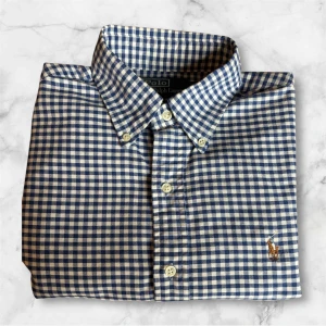 Blårutig skjorta från Polo Ralph Lauren - Snygg blårutig skjorta från Polo Ralph Lauren med klassisk button-down krage och broderad logga på bröstet. Skjortan har långa ärmar och vita knappar framtill. Perfekt för en stilren look.