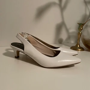 Vita pumps med klack och spetsig tå - Säljer ett par stilrena vita pumps med öppen häl och spetsig tå. Skorna har en medelhög klack och är tillverkade i ett slätt material som ger en elegant look. Perfekta för dig som vill ha en klassisk och trendig sko.