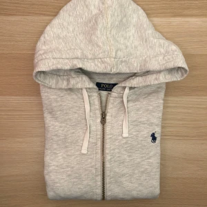 Ralph Lauren Hoodie Grå S - Ljusgrå hoodie med dragkedja från Polo Ralph Lauren. Tröjan har huva med dragsko, två fickor framtill och en broderad marinblå logga på bröstet. Tillverkad i mjuk bomullsblandning med polyester för extra komfort.