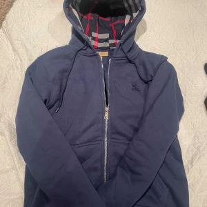 Navy blue zip-hoodie från Burberry - Mörkblå hoodie med dragkedja från Burberry. Klassisk rutig huva i rött, vitt och svart. Broderad logga på bröstet och justerbar huva med snörning. Perfekt för en avslappnad stil. Skick 9/10. Storlek S. Pris kan diskuteras!