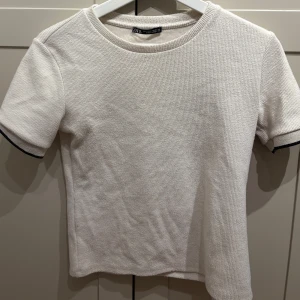 Vit ribbad t-shirt från Zara - Vit ribbad t-shirt från Zara med korta ärmar och svart kant på ärmsluten. Enkel och stilren modell med rund halsringning, perfekt för en clean look.