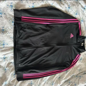 Svart träningsjacka med rosa detaljer från Adidas - Säljer en svart träningsjacka från Adidas med rosa ränder längs ärmarna och rosa logga på bröstet. Jackan har dragkedja framtill och är långärmad. Perfekt för träning eller sportiga outfits.