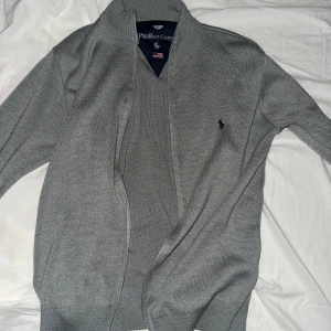 Grå kofta från Polo Ralph Lauren - Säljer en klassisk grå kofta från Polo Ralph Lauren med diskret broderad logga på bröstet. Koftan har lång ärm och är tillverkad i ett mjukt material som passar perfekt till lager-på-lager-stil.helt ny nästan 