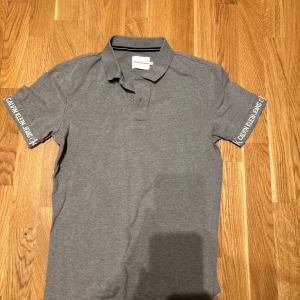 Grå piké från Calvin Klein Jeans - Säljer en stilren grå pikétröja från Calvin Klein Jeans med klassisk krage och korta ärmar. Logotypen finns tryckt på båda ärmarna. Perfekt för en clean och avslappnad look.