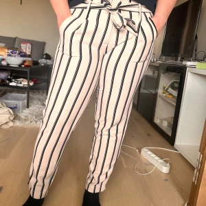 Randiga byxor med knytband i midjan - Snygga vita byxor med svarta vertikala ränder och knytband i midjan. Byxorna har normal passform, raka ben och sidofickor. Perfekta för en trendig och avslappnad look.