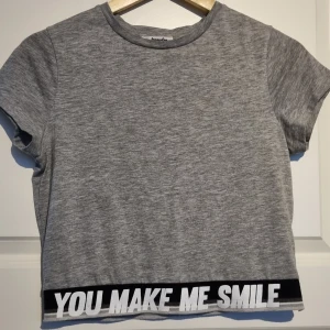 Grå croppad t-shirt från Jennyfer - Grå t-shirt från Jennyfer med korta ärmar och svart kant nertill med texten 'YOU MAKE ME SMILE' i vitt. Modellen är croppad och har en enkel, sportig stil. Perfekt till jeans eller shorts.