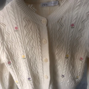 Beige stickad kofta med broderade blommor från Zara - Superfin beige stickad kofta från Zara med broderade små blommor i rosa, gul och lila. Koftan har vackert mönsterstickat tyg, rund halsringning och stora knappar framtill. Perfekt att slänga över en topp för en söt look.