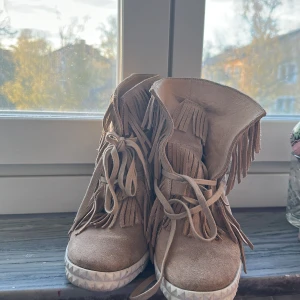 Beiga kilklackssneakers med fransar - Säljer ett par beiga sneakers med kilklack och snygga fransdetaljer. Skorna har snörning framtill och är gjorda i mocka. Perfekta för dig som vill ha en unik och trendig look.dom har lite fläckar på sig men inget märkvärdigt tycker jag, speciellt om man har Jens till !💕