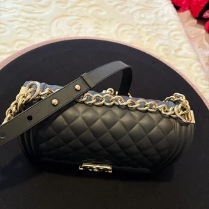 Svart quiltad axelväska. - Säljer en svart quiltad axelväska med kedjerem i guld och läder. Klassisk rektangulär form med elegant lås framtill och lyxig känsla. Perfekt för dig som vill ha en tidlös och trendig accessoar.