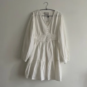 Vit linnéklänning från Neo Noir - Superfin vit klänning i linne från Neo Noir, modell Rihana Linen Dress. Klänningen har v-ringning, knappar framtill, markerad midja och långa ärmar. Perfekt för sommarens alla tillfällen!
