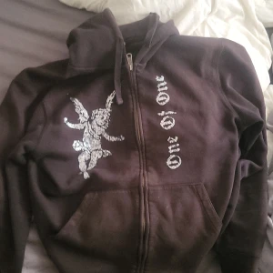 Svart hoodie med ängeltryck från One of One - Svart hoodie med dragkedja och huva från One of One. På framsidan finns ett stort vitt tryck av en ängel och texten 'One of One'. Hoodien har fickor framtill och ribbade muddar.