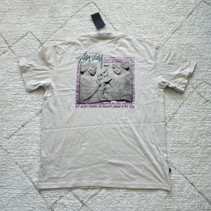 Beige t-shirt från Stüssy med grafiskt tryck - Säljer en beige t-shirt från Stüssy med grafiskt tryck på ryggen och liten logga framtill. T-shirten har normal passform och är tillverkad i mjuk bomull. Perfekt för dig som gillar streetwear och snygga detaljer.