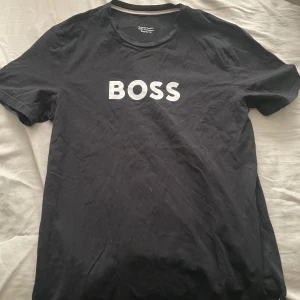 Svart t-shirt med BOSS-tryck - Säljer en svart t-shirt från BOSS med vitt tryck på bröstet. Klassisk rund halsringning och normal passform. Perfekt för en stilren och enkel look.