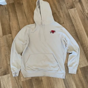 Beige hoodie med broderat hjärta - Säljer en beige hoodie med huva och magficka. På bröstet finns ett broderat rött hjärta med vingar. Tröjan har långa ärmar och ribbade muddar. Perfekt för en avslappnad stil.