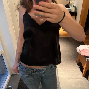 Svart satinlinne med smala axelband - Säljer ett svart linne i satin med smala axelband och v-ringning. Snyggt och enkelt plagg som passar till mycket. Materialet är glansigt och mjukt, vilket ger en lyxig känsla.