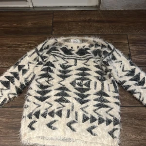 Vit och svart fluffig tröja med mönster - Säljer en supermjuk och fluffig tröja i vitt med svart grafiskt mönster. Tröjan har lång ärm och en avslappnad passform, perfekt för kyliga dagar. Ger en cool och trendig look till din outfit!