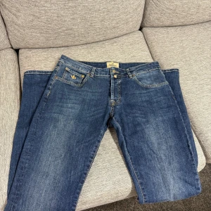 Blå jeans Morris Jeans 33/34 - Klassiska blå jeans från Steve Jeans med guldfärgad broderad detalj på myntfickan. Jeansen har normal passform och raka ben, perfekt för en avslappnad stil. Gylf med dragkedja och knäppning framtill.