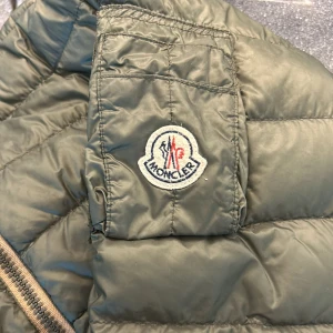 Moncler jacka - Säljer en grön pufferjacka från Moncler med orange foder. Jackan har lång ärm, dragkedja framtill och ribbade muddar vid ärmslut och krage. Klassisk Moncler-logga på ärmen. Perfekt för kyligare dagar. Jackan har en lagning på höger axel, kan ses på sista bilden, annars är den i bra skick. Passar för dig som är runt 155-160. Kom med prisförslag!😁