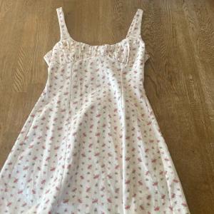 Vit blommig klänning från Zara - Söt vit klänning från Zara med små rosa blommor och breda axelband. Klänningen har en rynkad överdel och lätt utställd kjol, perfekt för varma dagar. Materialet känns mjukt och luftigt.