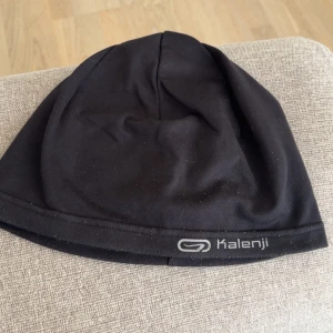 Svart mössa från Kalenji - En svart, tunn och smidig mössa från Kalenji. Perfekt för löpning eller träning utomhus. Tillverkad i syntetmaterial som andas och sitter bekvämt på huvudet. Diskret logga framtill.