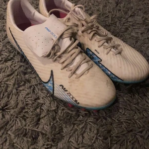 Nike Air Zoom fotbollsskor i vitt och blått - Säljer ett par Nike Air Zoom fotbollsskor i vitt med blå detaljer och svart Nike-logga. Skorna har snörning och mönstrad sula med rosa och svarta dobbar. Perfekta för dig som vill ha snygga och funktionella skor på planen.