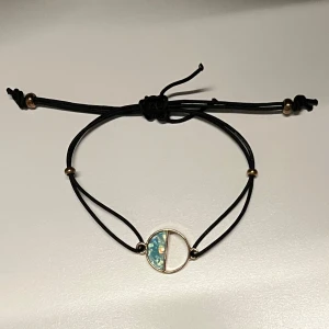 Svart armband med guld- och turkosdetalj i cirkel - Snyggt svart armband med justerbart band och en rund guldtonad detalj i mitten, dekorerad med turkosa inslag. Bra för sommar och strand. Troligen köpt på semester. 