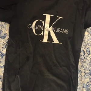 Svart t-shirt från Calvin Klein Jeans - Svart t-shirt med stort vitt CK-tryck och texten Calvin Klein Jeans på bröstet. Klassisk passform och rund halsringning. Perfekt för en stilren och enkel look.