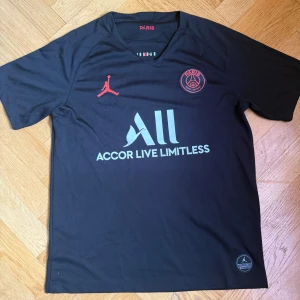 Svart PSG fotbollströja med röda detaljer - Säljer en svart PSG fotbollströja med röda detaljer och Jordan-logga. Tröjan har korta ärmar, rund hals och klubbmärke på bröstet. Materialet är lätt och andas, perfekt för fotboll eller träning.