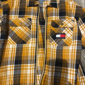 Gul och svart rutig overshirt från Tommy Jeans - Säljer en snygg overshirt från Tommy Jeans i gult, svart och vitt rutigt mönster. Skjortan har två bröstfickor med knappar och klassisk krage. Perfekt att bära som lager över en t-shirt. Använd max 3 gånger