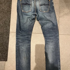 Blå jeans från Nudie Jeans - Säljer ett par klassiska blå jeans från Nudie Jeans med snygga slitningar och kontrastsömmar. Modellen har raka ben och femficksdesign med tydliga bakfickor. Perfekta för dig som gillar en tidlös jeanslook. Storlek 29/32 men sitter mer som 30 i midjan