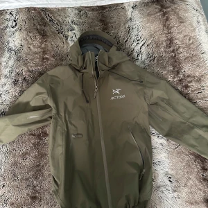 Olivgrön skaljacka från Arc'teryx - Säljer en olivgrön skaljacka från Arc'teryx med huva och dragkedja framtill. Jackan har två sidofickor med dragkedja och justerbar huva. Perfekt för utomhusaktiviteter och skyddar mot vind och regn. Använd 1-2 gånger.