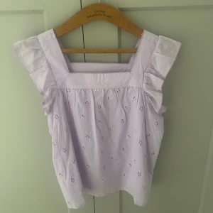 Ljuslila blus med volangärmar - Söt ljuslila blus med broderade blommor och volangdetaljer vid axlarna. Blusen har fyrkantig ringning och är ärmlös med breda axelband. Perfekt för varma dagar och ger en romantisk känsla.