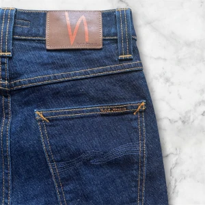 Grim trim nudie jeans - Säljer ett par klassiska mörkblå jeans från Nudie Jeans med raka ben och snygga kontrastsömmar. Byxorna har fem fickor, knapp och dragkedja i gylfen samt läderpatch med logga bak i midjan.